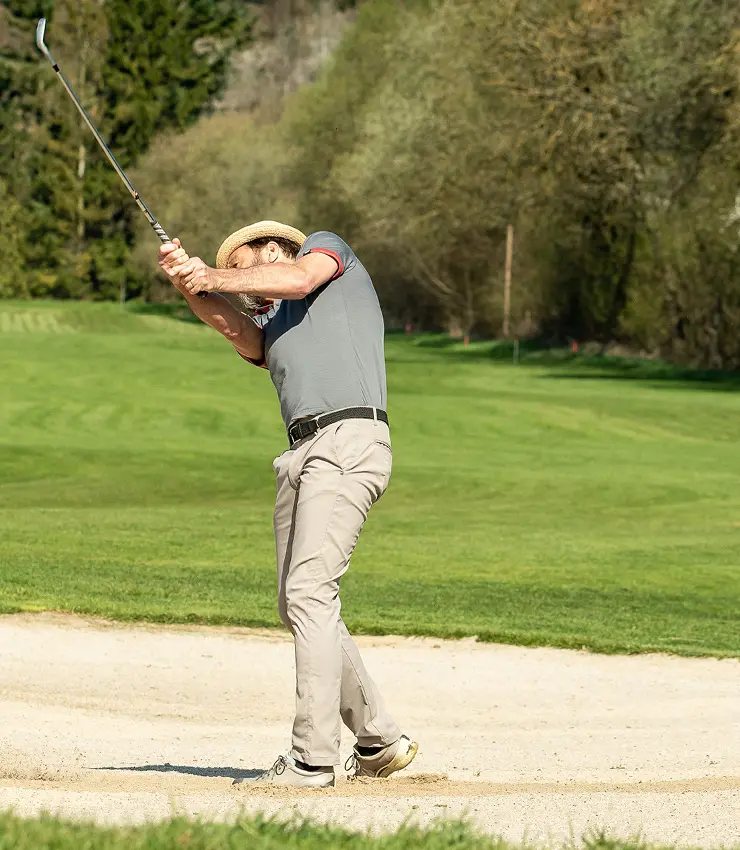 mensch mit Hut schlägt Ball im Sandbunker ab